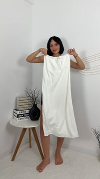 Biała sukienka do kolan z elastycznym paskiem w talii i detalem w postaci kokardy. Gładka tekstura, minimalistyczny design, prezentowana przez modelkę w pomieszczeniu.