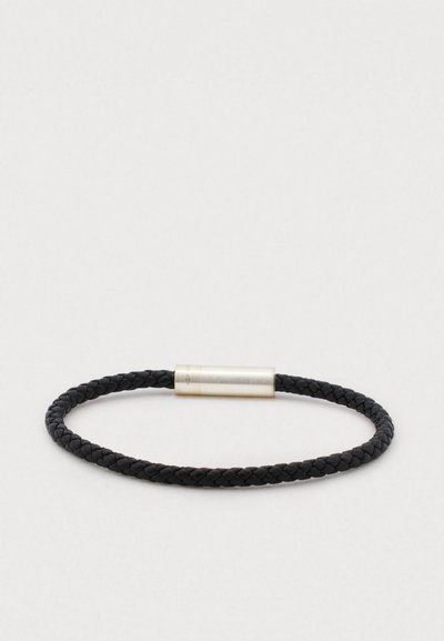 Fekete fonott bőr karkötő, henger alakú ezüst fém záróelemmel, sima textúrával, kerek formával, minimalista dizájn elemekkel.