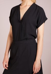 Kvinna i en kortärmad svart V-ringad blus som är instoppad i en högmidjad svart kjol med siddragkedja.