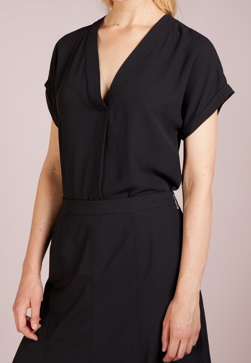 Kvinna i en kortärmad svart V-ringad blus som är instoppad i en högmidjad svart kjol med siddragkedja.