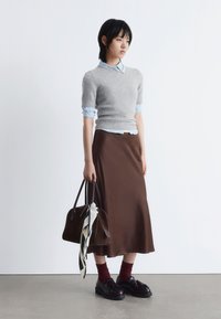 Model trägt einen grauen Kaschmirpullover über einem hellblauen Hemd, kombiniert mit einem langen braunen Satrock. Sie hält eine dunkelbraune Handtasche mit einem Schal.