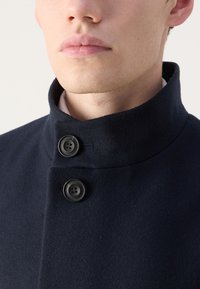 Cappotto di lana navy con colletto alto, dotato di due grandi bottoni neri e una texture liscia. Vista ravvicinata della parte superiore del capo.