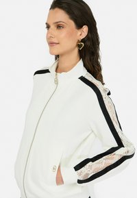 Veste blanche à zip, ornée de rayures noires et de dentelle sur les manches. Tissu texturé avec poches latérales et fermeture éclair en or.
