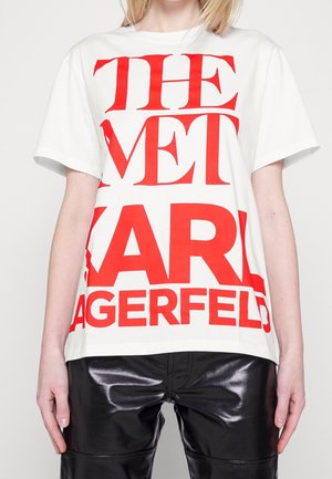 Osoba ubrana w białą koszulkę z dużym czerwonym napisem „THE MET KARL LAGERFELD” oraz czarne błyszczące spodnie, stojąca na jednolitym tle.