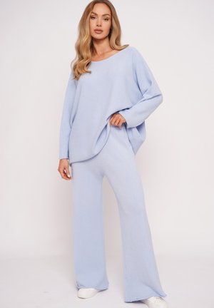 Kobieta ubrana w jasnoniebieski luźny sweter i pasujące szerokie spodnie oraz białe sneakersy, stojąca na jednolitym tle.