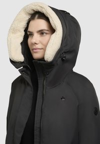 Schwarze Kapuzenjacke mit einem weichen, cremefarbenen Kunstfellfutter, Reißverschluss und Logo-Akzenten an der Brust und dem Ärmel. Strukturierter Schnitt und Material.