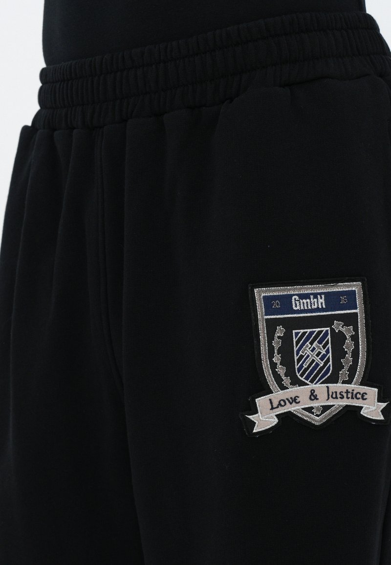 Black sweatpants with an elastic waistband and embroidered patch featuring a shield, text "GmbH," and "Love & Justice." Soft material, casual fit.