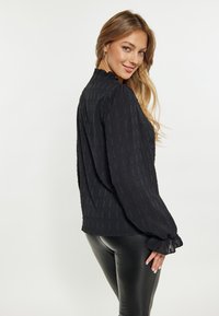Fekete blúz, amely érdes anyagból készült, függőleges vonalakkal, hosszú ujjakkal és fodros mandzsettával, fitt fekete leggings-szel párosítva.