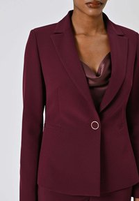 Blazer bordeaux con rever a lancia, chiusura con un bottone, due tasche laterali e vestibilità sartoriale. Sotto, strato marrone setoso visibile.