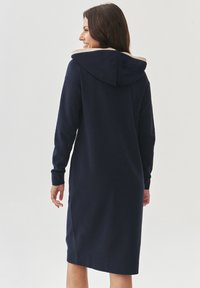 Navy hoodie jurk van zacht materiaal, voorzien van een beige gevoerde capuchon, lange mouwen en een rechte snit die tot halverwege de kuit valt.