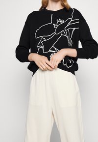 Czarny sweter z białym wzorem anatomicznego serca noszony z luźnymi białymi spodniami, ręce złożone na talii, na tle jasnym.
