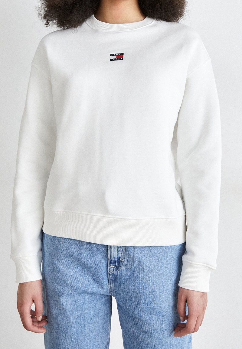 Persona che indossa una felpa bianca Tommy Jeans e jeans azzurri chiari, in piedi contro uno sfondo semplice, mostrando la parte superiore del corpo e le mani.