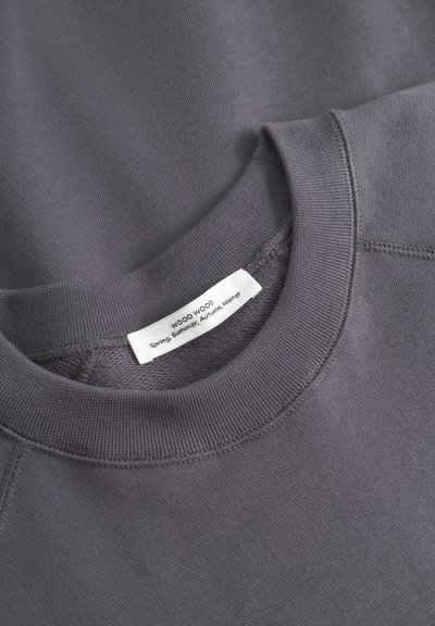 Grauer Sweatshirt mit rundem, geripptem Halsausschnitt, strukturiertem Stoff und einem Etikett mit "WOOD WOOD" und saisonalen Details.