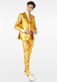 Completo metallico oro con giacca aderente e pantaloni affusolati, abbinato a una camicia bianca e una cravatta dorata. Sneakers bianche completano il look.