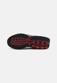 Semelle d'une chaussure de sport noire et rouge foncé montrant un motif de la semelle texturé avec des accents rouges circulaires et un petit logo swoosh noir.