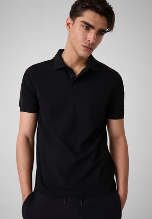 Junger Mann mit kurzen dunklen Haaren, der ein schwarzes kurzärmeliges Poloshirt trägt, steht mit den Händen in den Taschen vor einem schlichten hellgrauen Hintergrund.