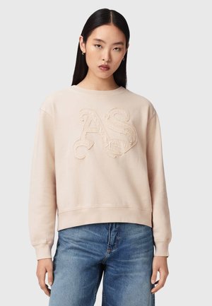 Kobieta z długimi ciemnymi włosami, ubrana w beżową bluzę z teksturowanymi literami "AS" oraz niebieskie dżinsy, stojąca na jednolitym tle.