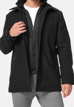Homme portant un manteau en laine foncée par-dessus une veste matelassée noire zippée et un t-shirt blanc, avec un pantalon gris, visible du cou jusqu'à mi-cuisse.