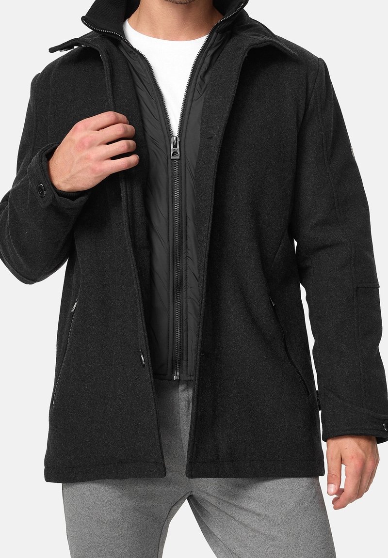 Homme portant un manteau en laine foncée par-dessus une veste matelassée noire zippée et un t-shirt blanc, avec un pantalon gris, visible du cou jusqu'à mi-cuisse.
