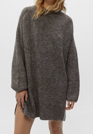 Femme portant une robe pull tricotée ample à manches longues gris foncé avec une fente latérale, debout devant un fond blanc uni.