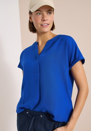 Femme portant une casquette beige, un chemisier bleu royal à manches courtes et un pantalon bleu marine foncé à cordon, regardant légèrement sur le côté et souriant.