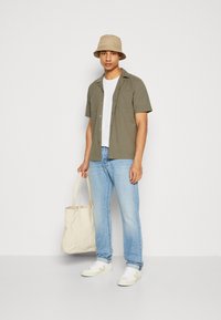 Camisa de manga corta verde olivo con dos bolsillos, camiseta blanca, jeans de mezclilla azul claro, gorra de cubo beige y bolso de crema con acentos claros.