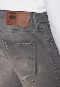 Jeans in denim grigio con dettagli strappati, un'etichetta in pelle marrone con logo e struttura dei taschini standard.