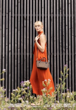 Woman in a sleeveless orange dress stands barefoot holding a woven handbag, against a tall vertical wooden slat background.