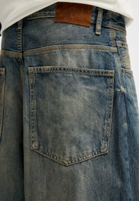 Pantalones cortos de mezclilla azul deslavada con una superficie texturizada, que cuentan con un parche de cuero marrón y dos bolsillos traseros con costuras en contraste.