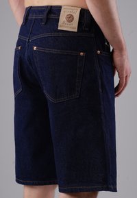 Denim shorts in dark blue with copper rivets, five pockets, and a tan leather patch on the back waistband featuring brand logo.