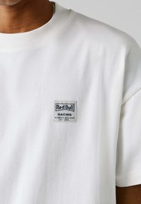 Λευκό βαμβακερό T-shirt με ριμπ λαιμόκοψη και μικρό γκρι σήμα που γράφει "Red Bull Racing Authentic Race Gear EST 2004."