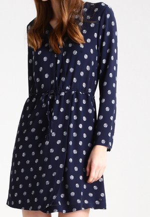 Granatowa sukienka z długim rękawem w białe groszki, ze sznurkiem w talii i zapinana na guziki, noszona przez kobietę o brązowych włosach.