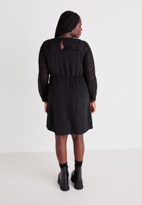 Robe noire avec des manches longues en dentelle, taille cintrée et détail au dos en forme de trou de serrure. Le tissu est lisse et présente une coupe décontractée.