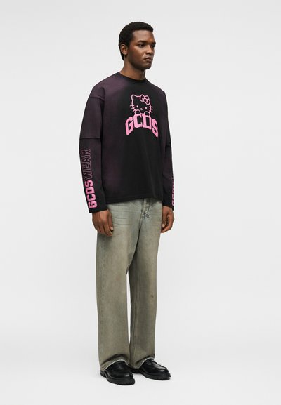 Άνδρας που φοράει μαύρο μακρυμάνικο πουκάμισο με ροζ λογότυπα Hello Kitty και GCDS, ξεθωριασμένο γκρι τζιν και μαύρα παπούτσια, στέκεται μπροστά σε λευκό φόντο.