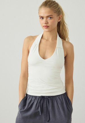 Young woman with blonde ponytail wearing white halter top and dark gray drawstring pants, standing against a plain light background.