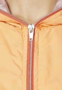 Giacca arancio chiaro con zip argentata, caratterizzata da un bordo corallo a contrasto lungo il cappuccio e la linea della zip. Tessuto dalla texture liscia.