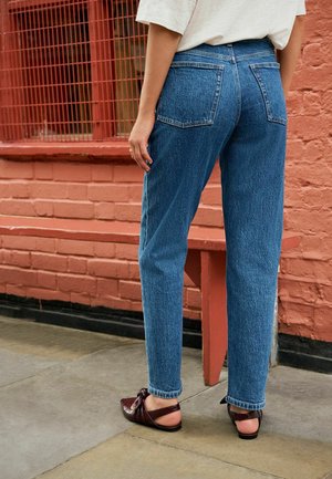 Jeans en denim bleu taille haute à coupe droite, dotés de poches arrières, d'un léger délavage et de bords légèrement effilochés, portés avec des mocassins bordeaux.