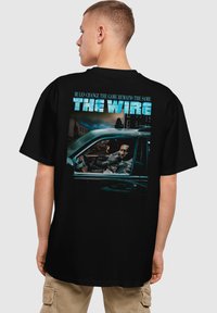 Svart T-skjorte med et grafisk motiv på baksiden som viser en bilscene, tekst som leser "THE WIRE", og et vintagebilde av karakterer innvendig.