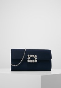 Navy modrá kabelka na ruku se stříbrnou květinovou přezkou s kamínky a stříbrným řetízkovým popruhem, umístěná na bílé podstavě proti šedému pozadí.