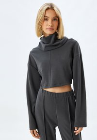 Ciemnoszara, skrócona bluza z wysokim kołnierzem, długimi rękawami i gładką fakturą. Noszona z dopasowanymi, ciemnoszarymi spodniami o wysokim stanie.
