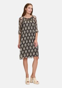 Schwarzes Kleid mit Blattmuster, transparenter Stoff, runder Ausschnitt und Dreiviertelärmel. Trägt beige Plateauschuhe.