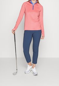 Růžová golfová košile s dlouhým rukávem a modrým zipem, spárovaná s navy úzkými kalhotami a bílými teniskami, držící golfovou hůl.