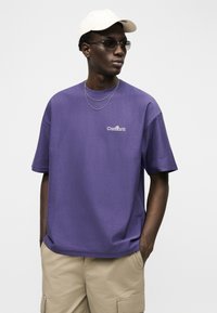 Άνδρας που φοράει μωβ μπλουζάκι Carhartt, μπεζ σορτς cargo, λευκό καπέλο, γυαλιά ηλίου και διακριτικά ασημένια κολιέ μπροστά σε απλό λευκό φόντο.