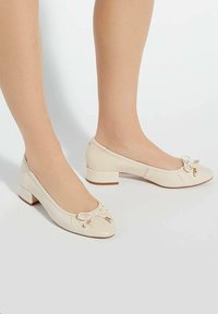 Chaussures à talons carrés en cuir beige avec un bout arrondi, ornées d'un petit nœud et d'accents dorés. Texture lisse avec des détails de design plissés.