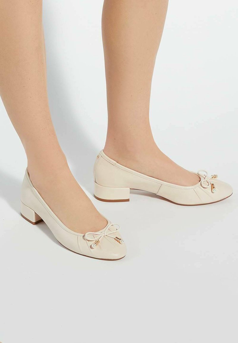 Chaussures à talons carrés en cuir beige avec un bout arrondi, ornées d'un petit nœud et d'accents dorés. Texture lisse avec des détails de design plissés.