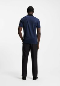 Dunkelblaues Poloshirt mit schwarzem Kragen, kurzen Ärmeln und subtiler Textur; kombiniert mit schwarzen Hosen und dunklem Schuhwerk.