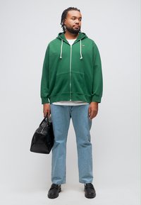 Άνδρας φορώντας πράσινο φούτερ με φερμουάρ, ανοιχτό μπλε τζιν και μαύρα παπούτσια, κρατώντας μία μαύρη τσάντα duffle με ουδέτερο φόντο.