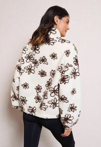 Fehér, bolyhos fleece dzseki barna virágmintával, magas gallérral, túlméretezett fazonban és rugalmas mandzsettával, fekete leggings felett viselve.