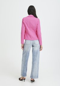 Eng anliegende pinke Jacke mit Kerbkragen, Rückenabnähern und langen Ärmeln. Kombiniert mit hellblauen, hoch taillierten Jeans und schwarzen Sandalen mit Absatz.