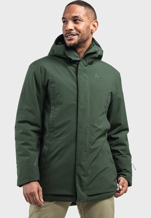 Mann trägt eine dunkelgrüne Kapuzen-Winterjacke mit Vordertaschen und beigen Hosen, steht vor einem schlichten hellen Hintergrund.